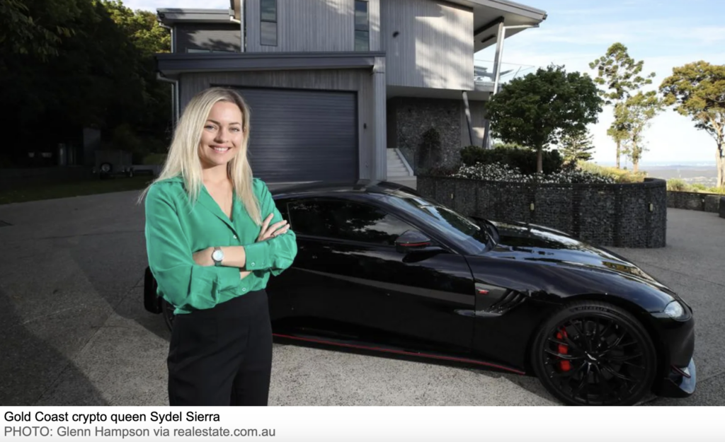 A woman with long blonde hair, wearing a green blouse and black pants, stands smiling with arms crossed in front of a sleek black sports car. Behind her is a modern multi-level house surrounded by greenery. The caption identifies her as Gold Coast crypto investor Sydel Sierra, known for helping others learn how to buy Bitcoin and crypto. 