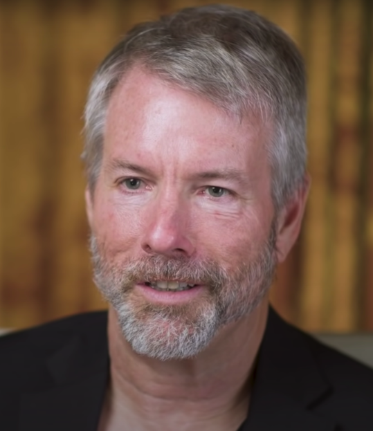Michael Saylor, co-founder and executive chairman of MicroStrategy, shown in a close-up portrait with grey hair and beard, wearing a dark blazer. Prominent figure in discussions about who owns most Bitcoin.